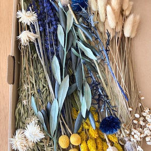 May include: A cardboard box filled with a variety of dried flowers and foliage. The arrangement includes lavender, olive branches, yellow billy buttons, blue thistle, and fluffy beige bunny tails. The flowers are arranged on a light brown surface.