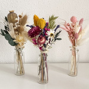 Könnte beinhalten: Drei kleine Glasvasen gefüllt mit Trockenblumenarrangements. Die Vase links enthält weiße und beige Trockenblumen, die mittlere Vase enthält lila, rosa und gelbe Trockenblumen und die Vase rechts enthält rosa und weiße Trockenblumen.