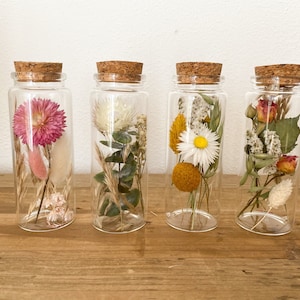 Könnte beinhalten: Vier Glasbehälter mit Korkdeckeln, gefüllt mit getrockneten Blumen. Die Behälter sind in einer Reihe auf einer Holzoberfläche angeordnet. Die Blumen umfassen Rosa, Weiß, Gelb und Rot.