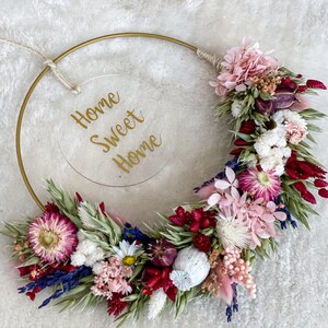 May include: A gold metal hoop wreath decorated with dried flowers in shades of pink, red, white, and blue. The wreath has a clear acrylic sign that reads "Home Sweet Home".