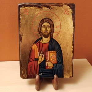 May include: Hand-painted religious icon depicting a figure with a halo, holding a book. The figure wears red and blue robes against a gold background. The icon is mounted on a wooden plaque and displayed on a wooden stand.