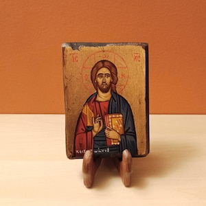 May include: A hand-painted wooden icon depicting Jesus Christ holding a book. The icon is painted in a traditional style with gold accents and the Greek text "IC XC NIKA" at the top.
