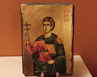 May include: A hand-painted icon of a saint holding a cross. The icon is on a wooden background with gold leaf accents. The saint is wearing a red and green robe and has a halo around his head. The text "ΘΕΟΥ ΔΟΥΛΟΣ" is written in Greek on the right side of the icon.
