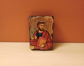 May include: A small wooden icon depicting a religious figure with a book in their hand. The icon is painted in a traditional style with gold accents and the text "Ο ΑΓΙΟΣ ΙΩΑΝΝΗΣ" at the bottom.