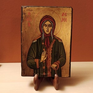 以下が含まれることがあります： 赤いスカーフと金の輪郭を持つ女性の、手描きのアイコン。アイコンは木製のスタンドに置かれ、赤い文字で「ΖΓ' ΝΗ」と書かれています。