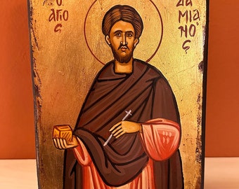 May include: A hand-painted icon of a saint holding a small golden box in their left hand and a cross in their right hand. The icon is painted on a wooden panel with a gold background and has Greek text in the top corners. The text reads "O Agios" and "Za Mia No G".