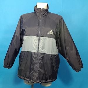 Op de afbeelding: Een zwart en grijs Adidas windjack met een volledige ritssluiting aan de voorzijde, een hoge kraag en lange mouwen met elastische boorden. Het jack heeft een horizontale grijze streep over de borst en het Adidas-logo in het wit.