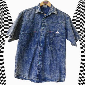 Può includere: Una camicia di jeans blu a maniche corte con un motivo bianco maculato. La camicia ha una chiusura frontale con bottoni, due tasche sul petto e un colletto a punta.