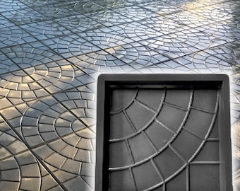 May include: A black, square paving stone mold with a fan-shaped design. The mold is next to a completed patio with a similar pattern. The patio stones are gray and arranged in a circular pattern.
