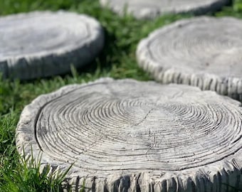 以下が含まれることがあります： 木の切り株の形をした灰色のコンクリート製の踏み石が4つあります。石は粗くて質感のある表面をしていて、緑の芝生の上に一列に並んでいます。