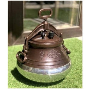 Op de afbeelding: Een bruine en zilveren snelkookpan met een handvat aan de bovenkant en zijhandvatten. De pan heeft decoratieve bloemen- en scriptdetails. De onderste helft is zilver en de bovenkant is bruin.