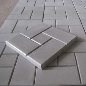 Op de afbeelding: Een grijze betonnen terrastegel met een uniek, in elkaar grijpend ontwerp. De tegel is gemaakt van een duurzaam materiaal en is perfect voor het creëren van een stijlvolle en functionele buitenruimte.