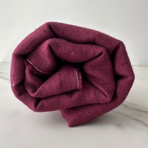 May include: Rolled-up burgundy fabric with a textured weave. The fabric is a deep, rich shade of red-purple, with visible white stitching along the edges. The material appears to be a medium-weight textile, suitable for various sewing projects.