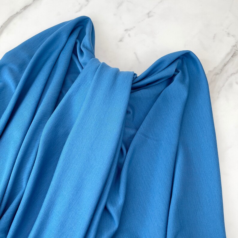 May include: A close-up of a blue fabric, folded and draped on a white marble surface. The fabric has a soft, smooth texture and a slightly textured appearance.