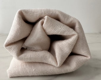 May include: A beige linen fabric rolled up into a cylinder shape. The fabric has a natural, textured look and feel.