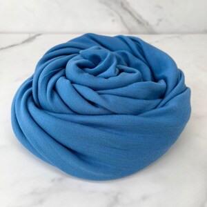 May include: A soft, blue fabric scarf is rolled up into a rose shape. The fabric is smooth and has a subtle sheen.