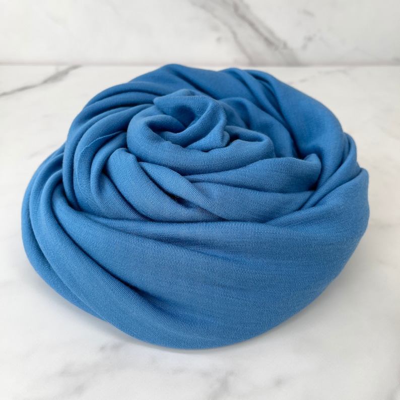 May include: A soft, blue fabric scarf is rolled up into a rose shape. The fabric is smooth and has a subtle sheen.
