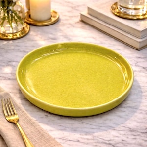 May include: A round, chartreuse-colored ceramic plate with a crackle glaze finish. A gold fork rests on a beige linen napkin. The plate is on a marble surface, with a candle and flowers in the background.