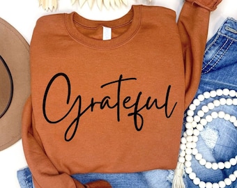 May include: A rust-coloured sweatshirt with the word "Grateful" written in black cursive lettering. The sweatshirt is laid out on a white surface with a brown hat, blue jeans, and a white beaded necklace.