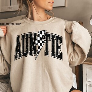 K&ouml;nnte beinhalten: Ein beigefarbenes Sweatshirt mit Rundhalsausschnitt und dem Wort AUNTIE in gro&szlig;en, schwarzen Blockbuchstaben mit Used-Optik. Der Buchstabe I ist durch einen schwarz-wei&szlig; karierten Blitz ersetzt. Der Text WILDLYFREETEESHOP ist in der Ecke zu sehen.