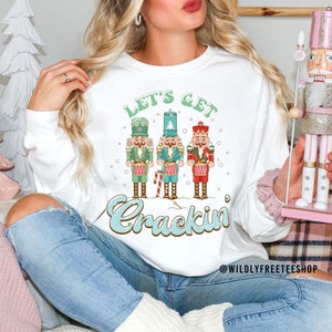 May include: A white long-sleeve sweatshirt featuring a graphic of three nutcrackers in green, blue, and red uniforms. The text above reads LET'S GET and below reads Crackin in blue script. The text WILDLYFREETEESHOP is printed in the bottom right corner.