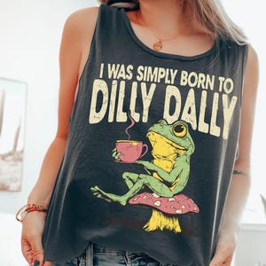Op de afbeelding: Donkergrijze tanktop met de tekst "I WAS SIMPLY BORN TO DILLY DALLY." De afbeelding toont een groene kikker op een paddenstoel, met een roze kopje. De kikker heeft gele accenten.