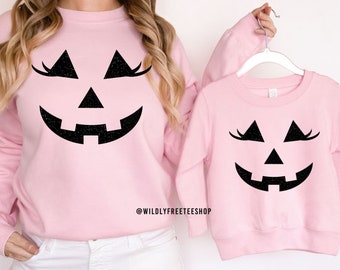 May include: Two pink sweatshirts with a black glitter jack-o'-lantern face with eyelashes. The text "@WILDLYFREETEESHOP" is on the bottom of the larger sweatshirt. The text "MADE WITH BLACK GLITTER" is at the bottom of the image.