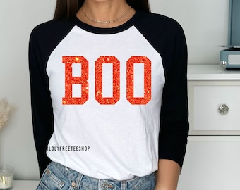Puede incluir: Una camiseta de béisbol blanca y negra con la palabra "BOO" en letras de purpurina naranja.