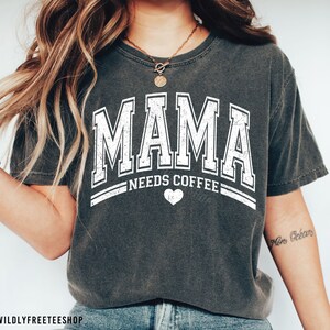 May include: A charcoal grey short-sleeved t-shirt featuring white distressed lettering that reads MAMA NEEDS COFFEE with a small heart icon centred below the text. The shirt has a crew neckline and a relaxed fit, showcasing a vintage-inspired graphic.