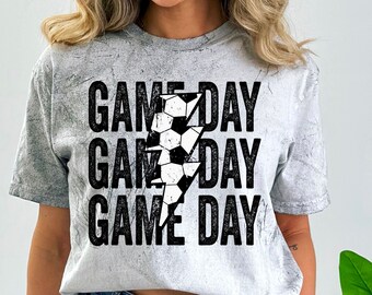 May include: A gray t-shirt with a distressed look featuring a black lightning bolt graphic with a soccer ball inside. The text "GAME DAY" is repeated three times in black.