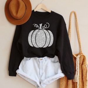 May include: A black sweatshirt featuring a silver glitter pumpkin design. The sweatshirt is worn over a pair of light blue denim shorts.