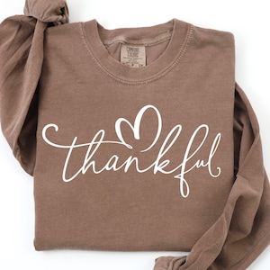May include: A long-sleeve crewneck sweatshirt in an espresso brown color featuring the word thankful in white cursive script with a heart shape above the letter k. The interior neck label displays the Comfort Colors brand name and size details.