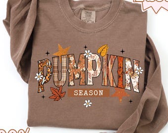 May include: A brown long-sleeved top with the words "PUMPKIN SEASON" in a seasonal design. The shirt is made of 100% cotton and has a crew neck. The design incorporates floral, plaid, and leaf patterns.