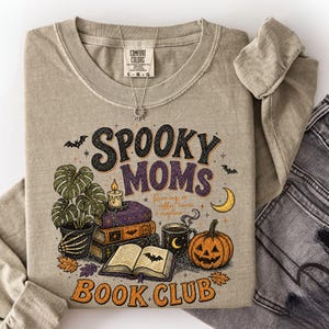 Puede incluir: Una camiseta beige de manga larga con un estampado gráfico que incluye el texto "SPOOKY MOMS BOOK CLUB" y "Running on coffee, books & chapters". El diseño muestra una planta en maceta, libros apilados, una vela, una calabaza y murciélagos.