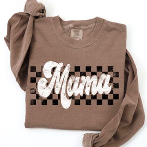 Puede incluir: Una camiseta de manga larga marr&oacute;n con la palabra "Mama" en una fuente blanca retro sobre un fondo a cuadros blanco y negro.