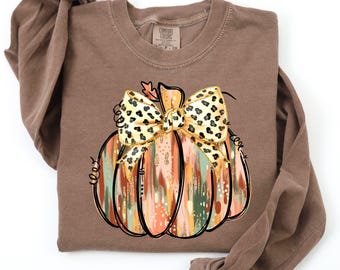 May include: A long-sleeve espresso brown shirt featuring a graphic of a pumpkin with a leopard print bow. The pumpkin has vertical brushstroke patterns in shades of orange, green, and cream. The shirt has a crew neckline and a Comfort Colors label.