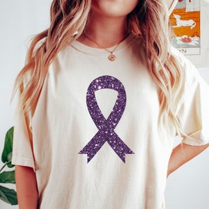 Puede incluir: Una camiseta blanca con un diseño de cinta de purpurina morada. La cinta es un símbolo de concienciación para muchas causas.