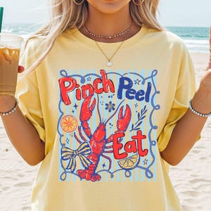Puede incluir: Camiseta amarilla clara con un diseño gráfico que presenta una langosta roja y las palabras "Pinch Peel Eat". El diseño incluye detalles en azul y naranja, ideal para un look casual y veraniego.