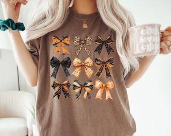 May include: A brown t-shirt with a graphic of multiple orange and black bows. The bows have different patterns, including polka dots, plaid, and leopard print.