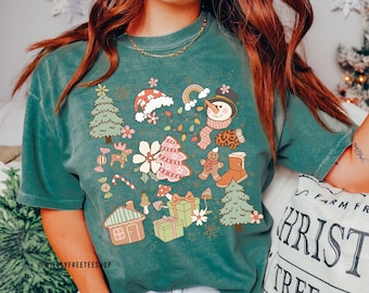 May include: A teal green t-shirt with a Christmas-themed design featuring a snowman, gingerbread man, candy canes, presents, and a gingerbread house. The design is in a vintage style with pastel colours.