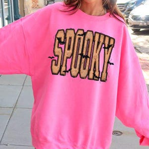 May include: A bright safety pink long-sleeve sweatshirt featuring the word SPOOKY printed in large, blocky, orange and black striped letters across the chest. The text is accented with small black bat silhouettes scattered around the letters.