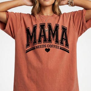 May include: A terracotta-coloured short-sleeve t-shirt featuring black distressed lettering that reads MAMA NEEDS COFFEE with a small heart icon centred below the text. The shirt has a relaxed fit and a crew neckline, showcasing a vintage-inspired graphic design printed on the front.