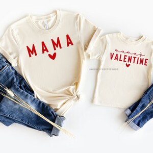May include: Two beige t-shirts with red text. The larger shirt says "MAMA" with a red heart. The smaller shirt says "mama's VALENTINE" with a red heart.