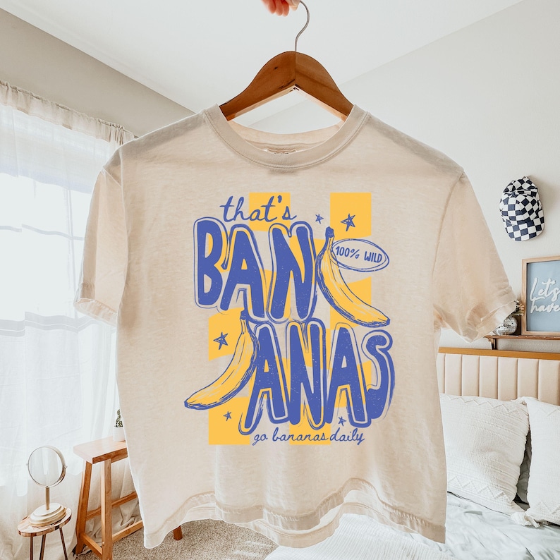 Pu&ograve; includere: T-shirt color crema con un design giocoso. Le parole "that's BANANAS" in blu su un quadrato giallo. Sono presenti anche due banane gialle e la frase "go bananas daily". La maglietta &egrave; appesa a una gruccia di legno.
