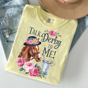 Może przedstawiać: Jasnoż&oacute;łta koszulka z napisem "Talk Derby to Me!" i grafiką konia w kapeluszu w kwiaty, koktajlem i r&oacute;żowymi r&oacute;żami. Koszulka leży na niebieskich dżinsach i beżowym kapeluszu.