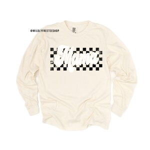 Puede incluir: Una camiseta de manga larga de color crema con un patr&oacute;n de cuadros negros y blancos y la palabra "Mama" en letras blancas.