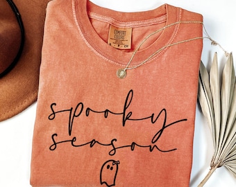 May include: A coral-coloured Comfort Colors t-shirt with the words "spooky season" in black script and a ghost graphic. A gold necklace with a pendant rests on the shirt. A brown hat and dried plant are in the background.