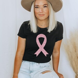 May include: A black short-sleeved t-shirt featuring a large, centred pink glitter awareness ribbon graphic. The shirt has a crew neckline and is paired with light blue denim jeans that have a distressed rip on the upper thigh area.