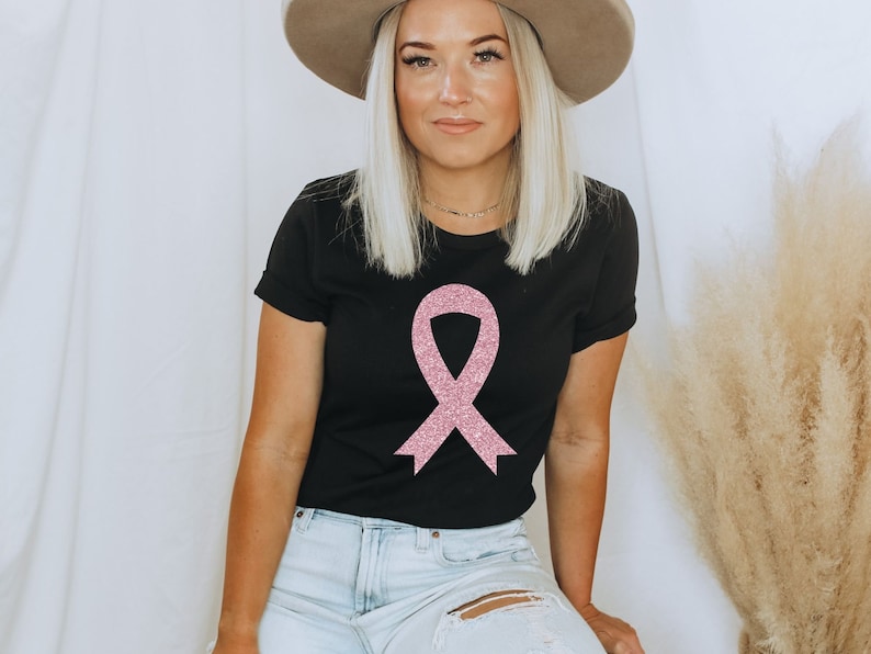 May include: A black short-sleeved t-shirt featuring a large, centred pink glitter awareness ribbon graphic. The shirt has a crew neckline and is paired with light blue denim jeans that have a distressed rip on the upper thigh area.