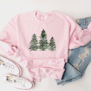 Puede incluir: Una sudadera rosa con un dise&ntilde;o de acuarela de tres &aacute;rboles verdes. La sudadera est&aacute; colocada sobre una superficie blanca con un par de zapatillas blancas y unos vaqueros azules.
